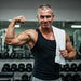 An older man with short dark grey hair lifting weights in a gym, wearing a black tank top, with gym equipment in the background.
